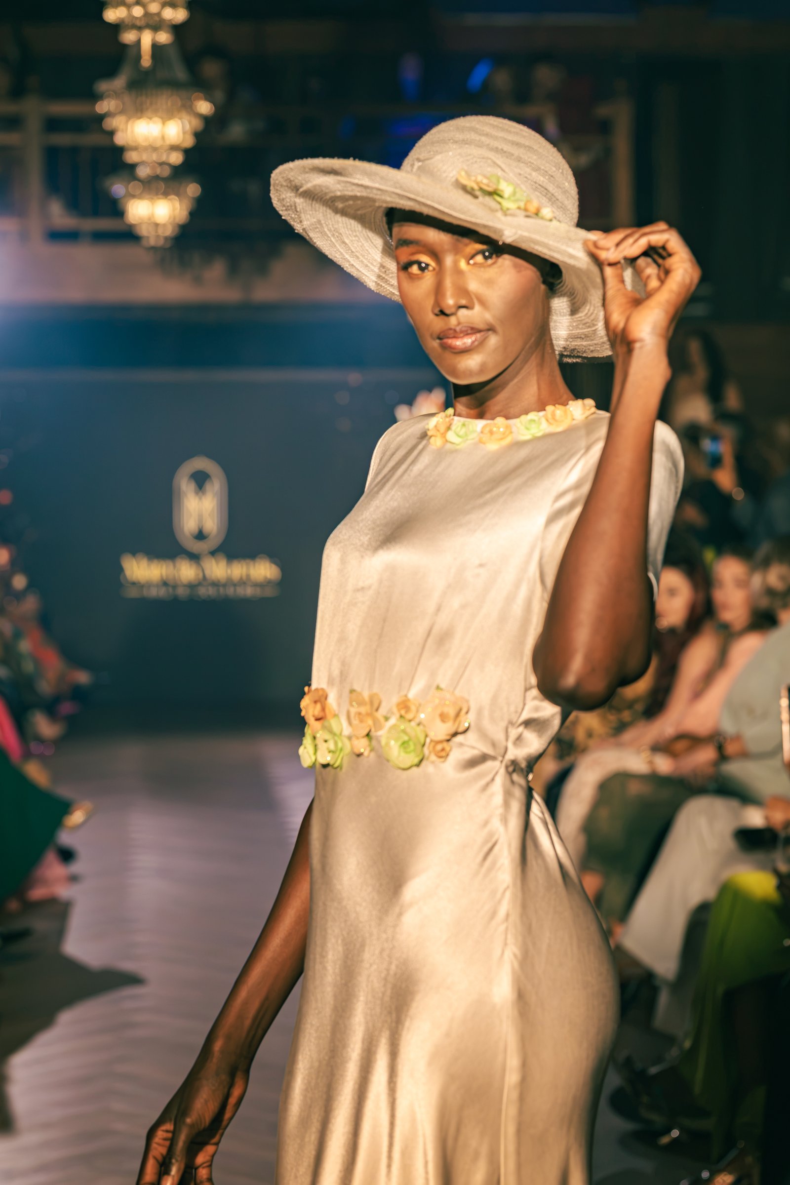 Runway model in a sleek silver silk dress with floral appliques and a matching wide-brimmed hat