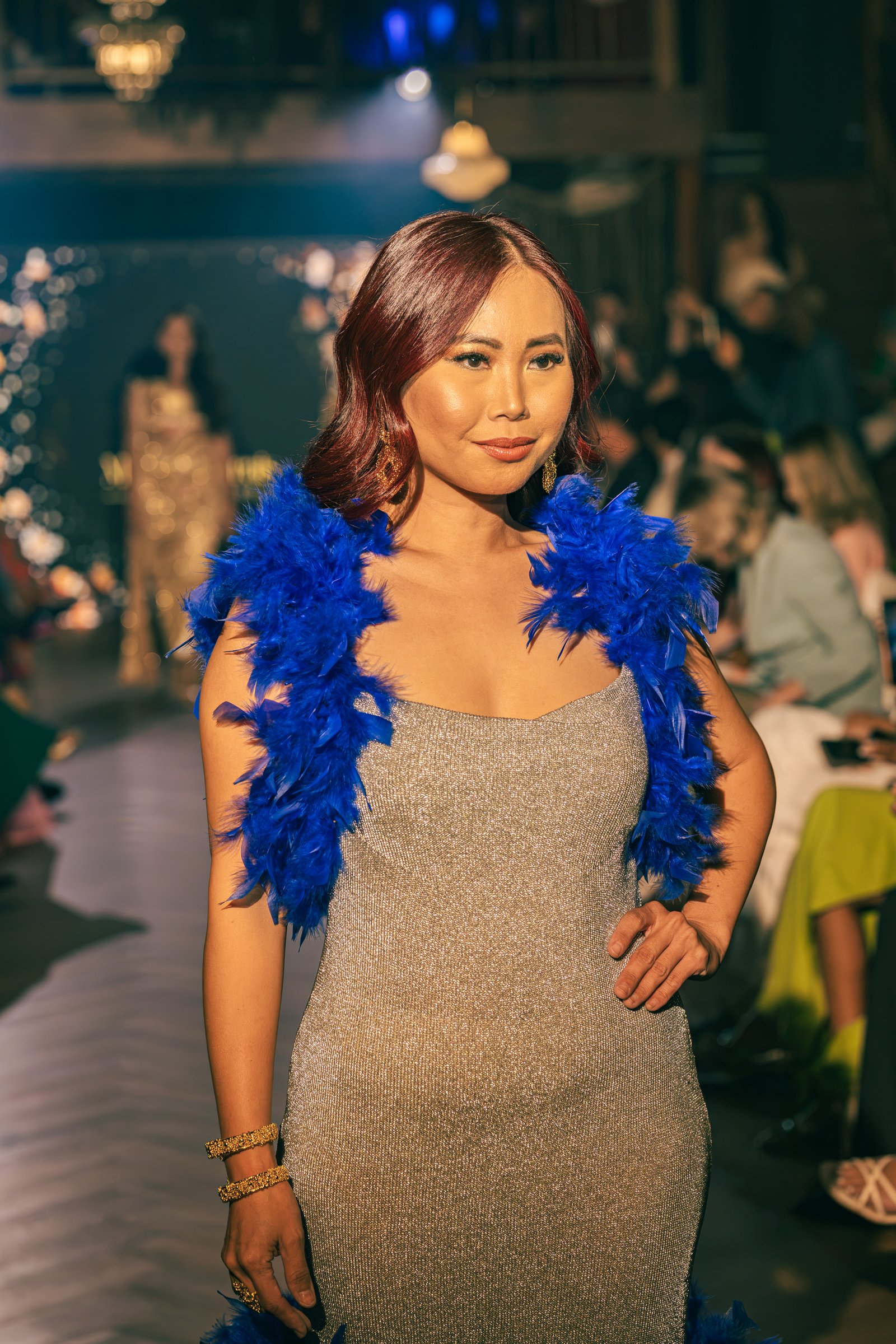 Runway model in a sparkling silver dress paired with a vibrant blue feather shoulder wrap